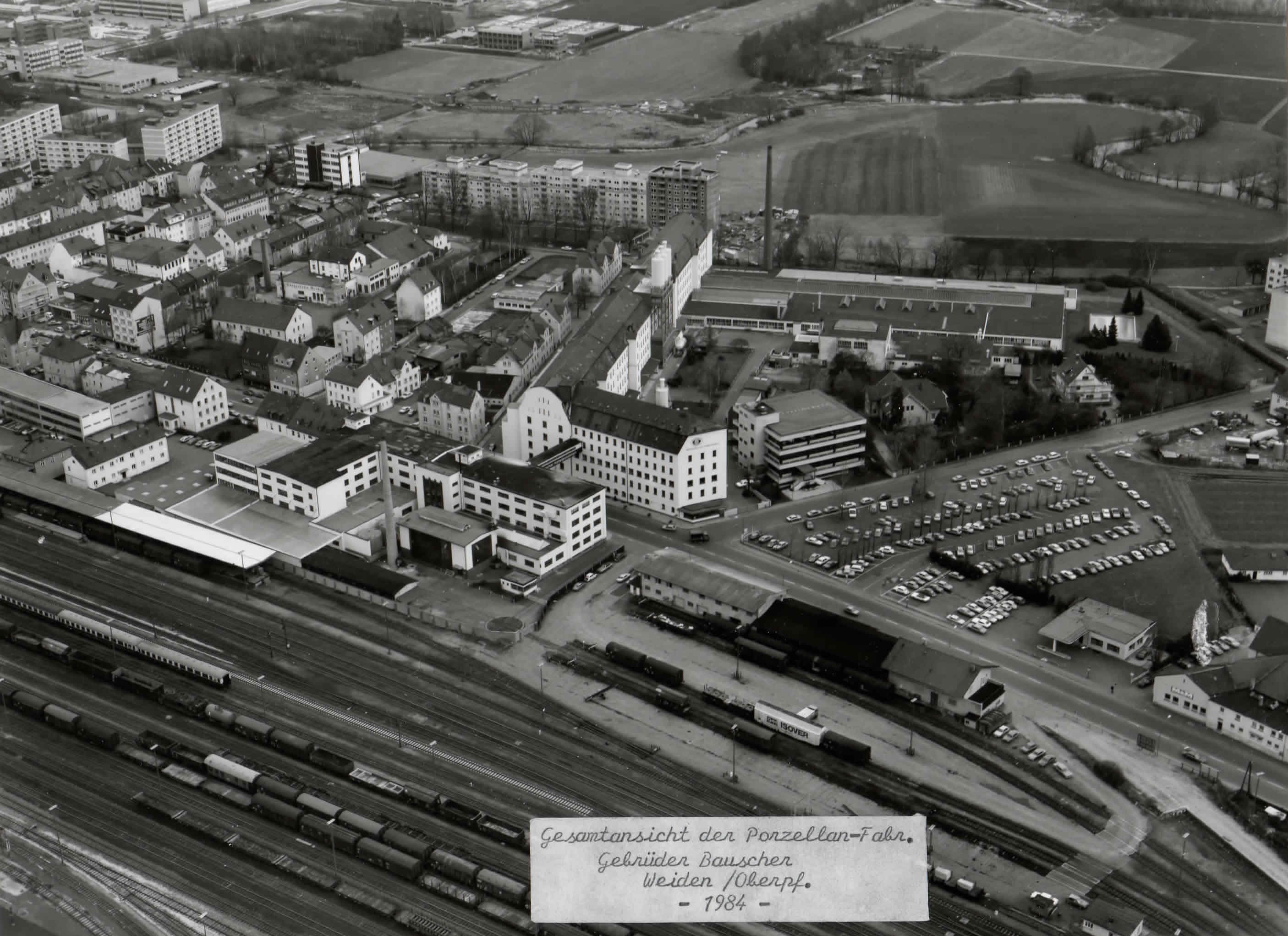 Schwarzweiß-Luftaufnahme der Bauscher-Fabrik mit angrenzenden Bahngleisen und der historischen Jahresbeschriftung 1984.