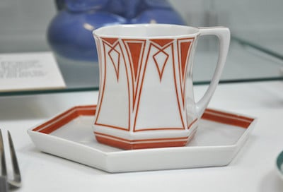 Kantige weiße Tasse mit rotbraunem geometrischem Dekor auf einer eckigen Untertasse.