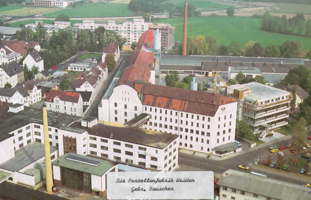 Farbige Luftaufnahme des Bauscher-Werks mit mehrgeschossigen Gebäuden, Schornstein und angrenzender Straße.