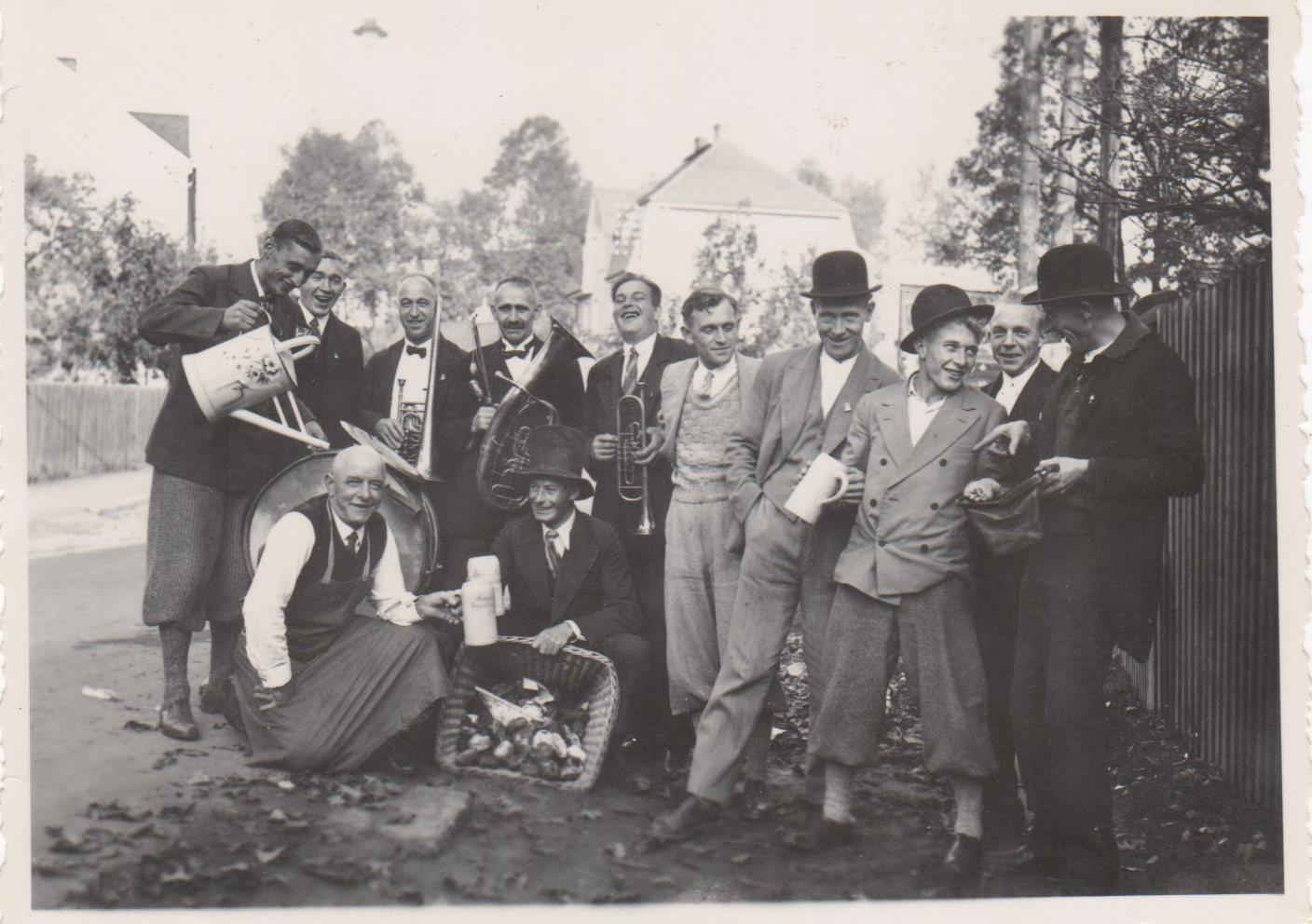 Festlich gekleidete Gruppe auf einer Dorfstraße mit Trinkgefäßen und einem Musikinstrument.