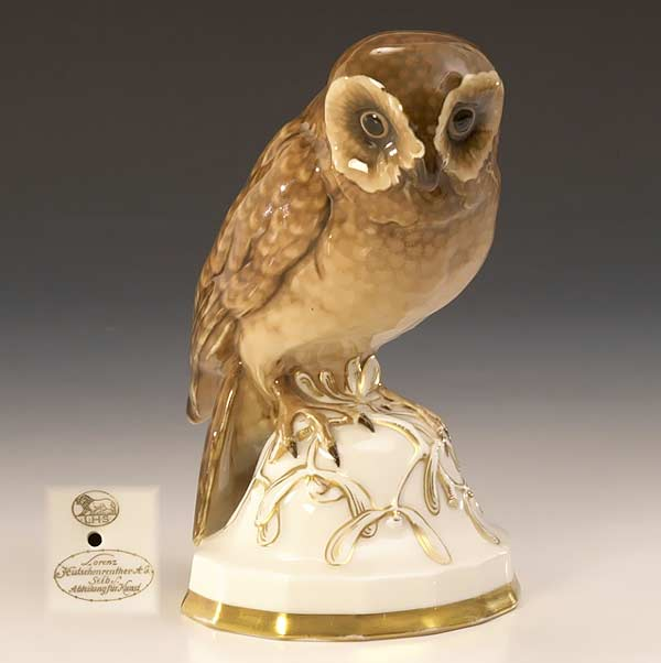 Braun und gold staffierte Porzellaneule auf hellem, ornamentiertem Sockel; unten links eine kleine Markendarstellung.