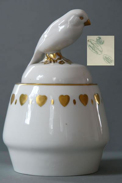 Kleine weiße Deckeldose mit aufgesetztem Vogel und einer Reihe goldfarbener Ornamente.