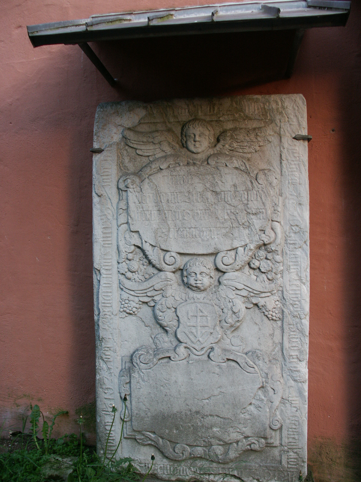 Grabplatte an der Kirchenwand. Engelsköpfe, Wappen, Fruchtgehänge und Schriftfelder schmücken den Stein.