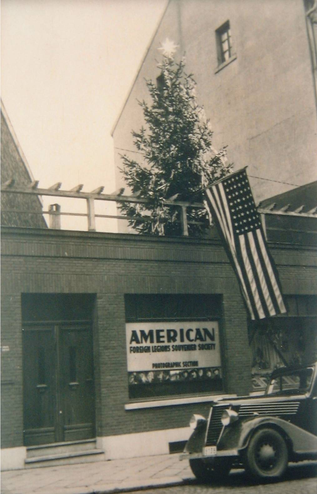 Historische Straßenaufnahme eines Fotogeschäfts in Eupen mit amerikanischer Flagge und parkendem Fahrzeug.