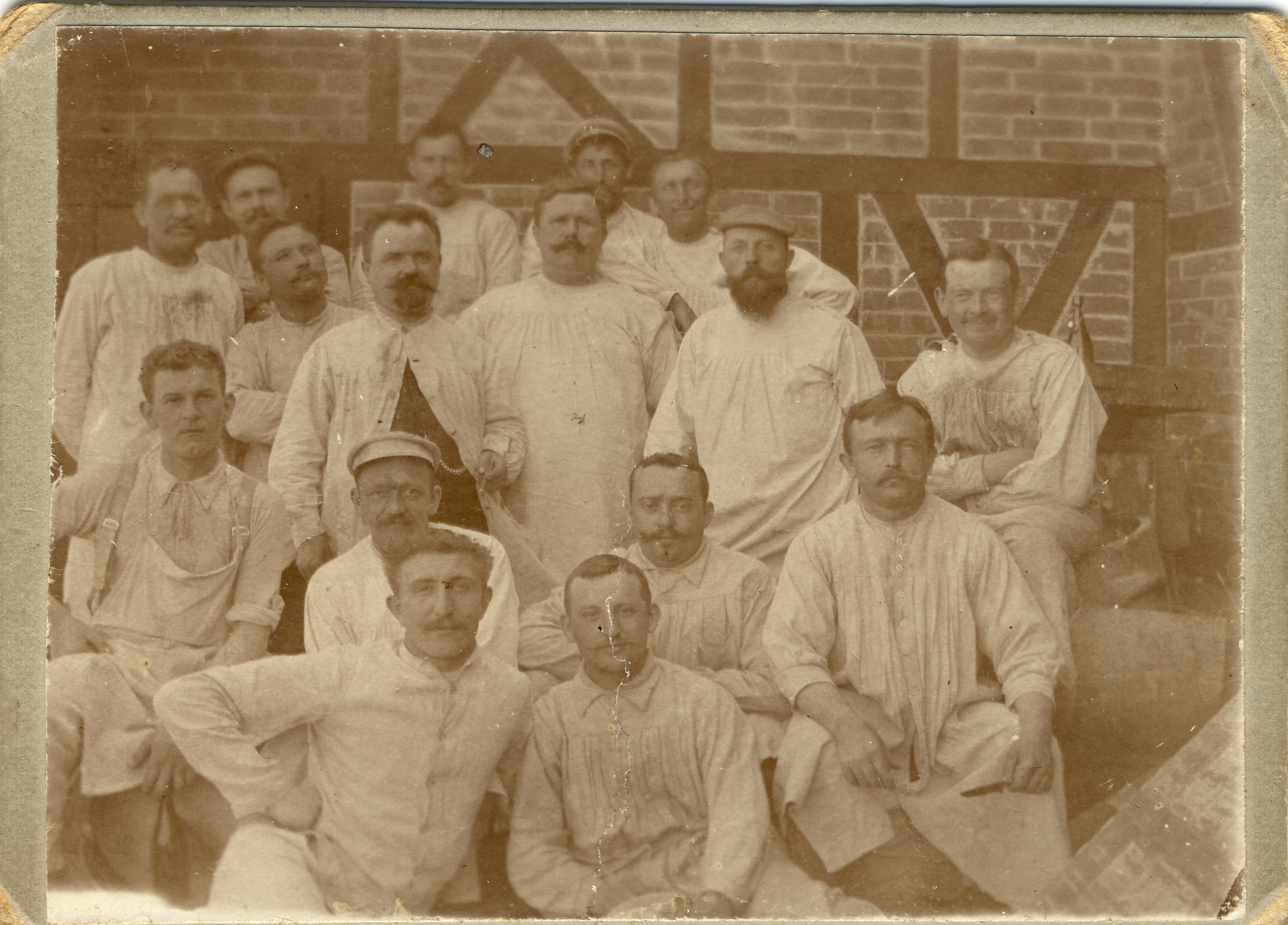Historische Gruppenaufnahme von Männern in Arbeitskitteln vor einer Backsteinwand der im Fundus Roesler zugeordneten Fabrik.