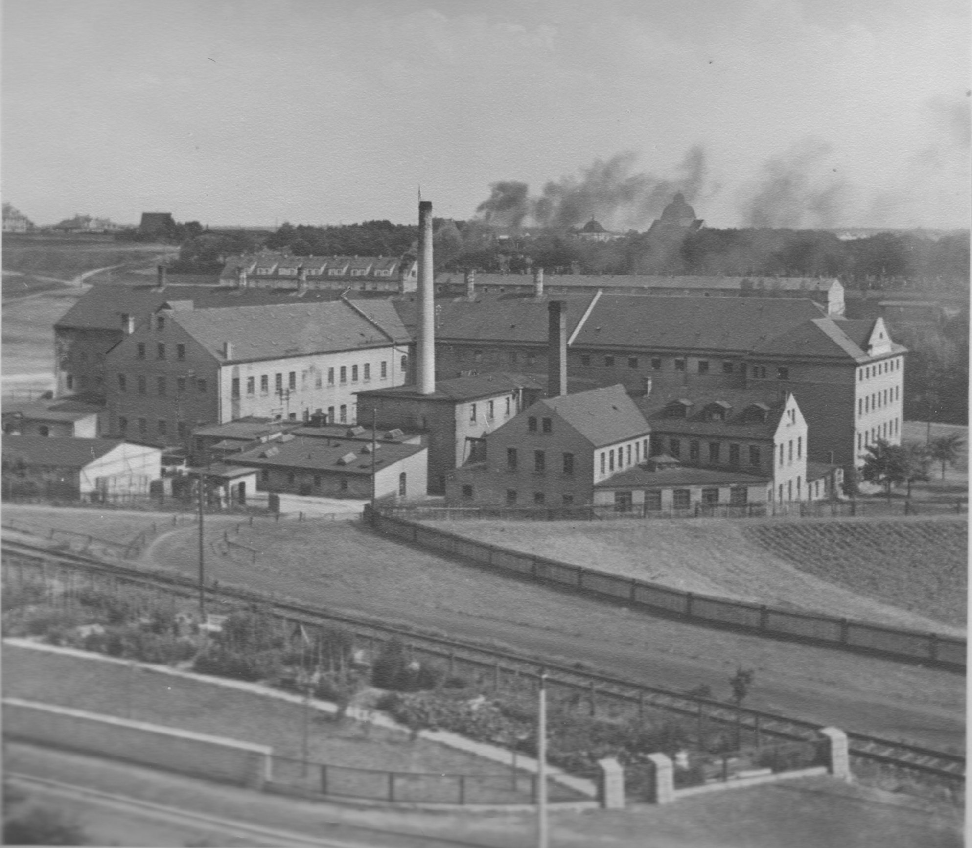 Fabrikanlage mit Schornsteinen, mehrgeschossigen Gebäuden und Gleisen im Vordergrund.
