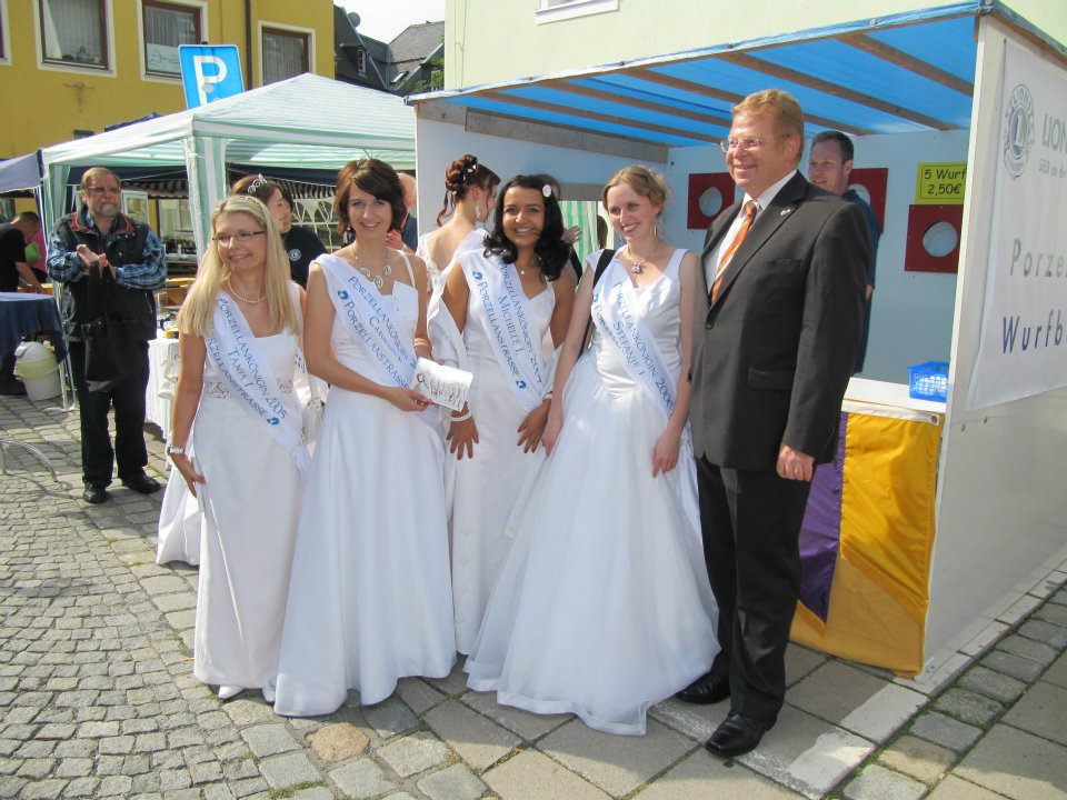 Mehrere Frauen mit Porzellanstraßen-Schärpen und ein Begleiter vor einem Marktstand mit porzellanbezogener Beschriftung.