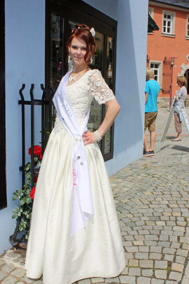 Ganzaufnahme eines festlichen hellen Kleides mit Schärpe vor einem Gebäude.