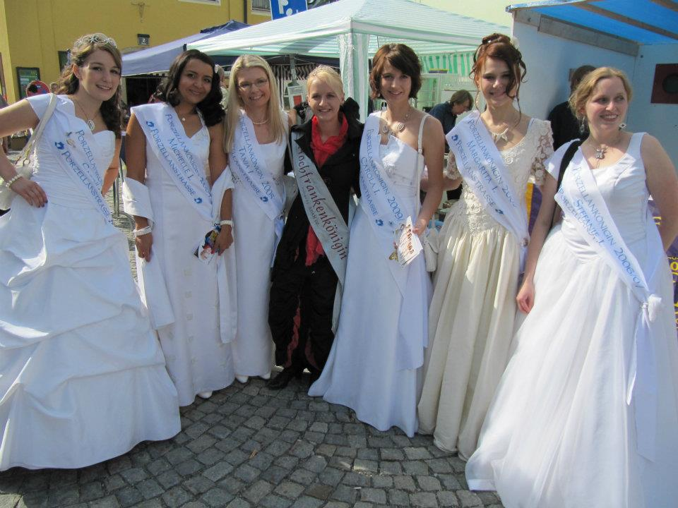 Mehrere Frauen in hellen Festkleidern und mit Porzellanstraßen-Schärpen gemeinsam mit einer rot gekleideten Begleiterin vor einem Marktstand.