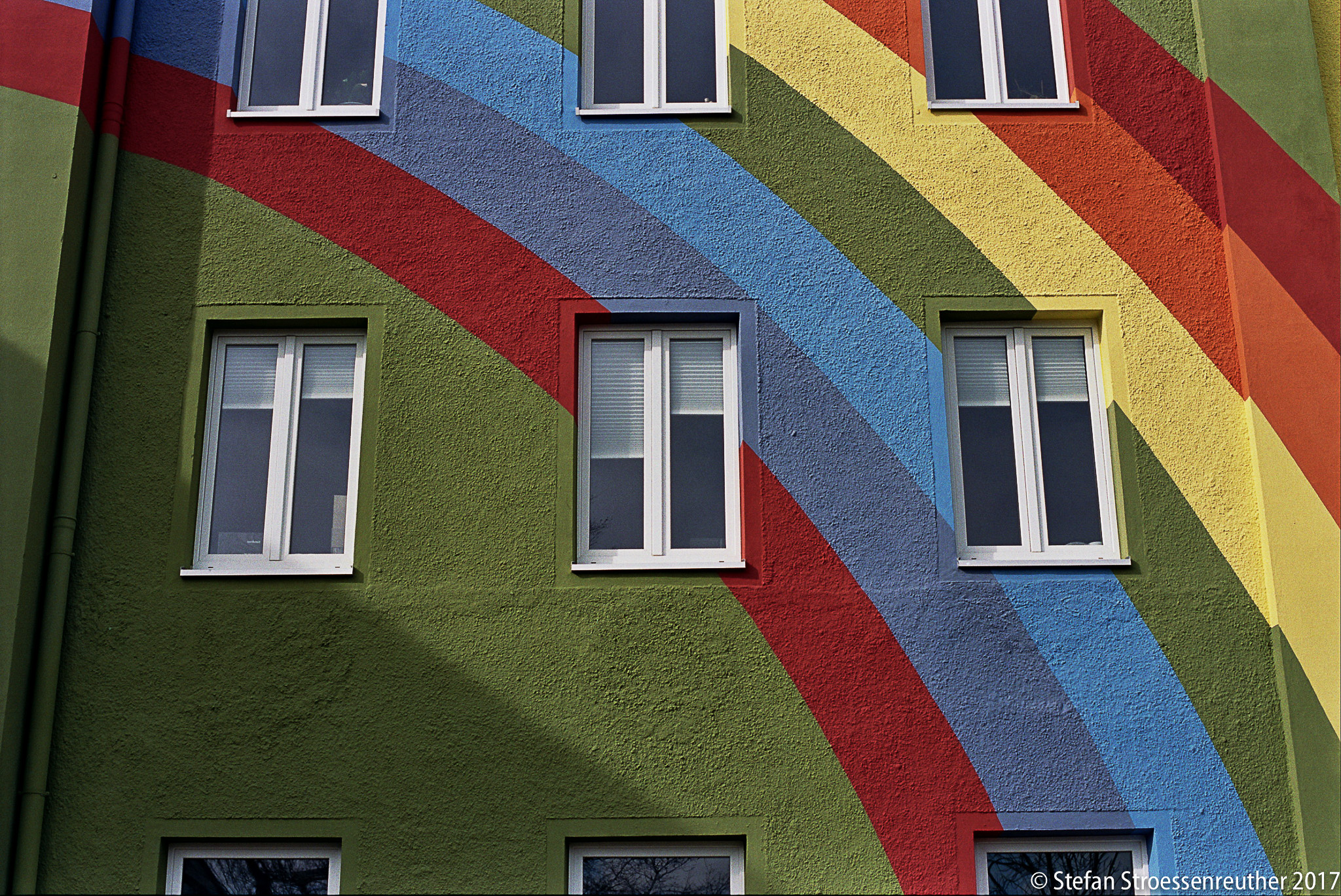 Detail der Regenbogenfassade mit roten, gelben, grünen und blauen Bändern zwischen weißen Fensterrahmen.