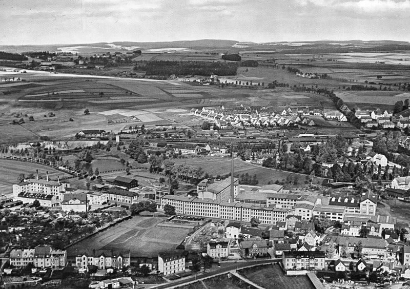Historische Luftaufnahme mit einem großen Fabrikkomplex, Wohnbebauung und landwirtschaftlich genutzten Flächen.