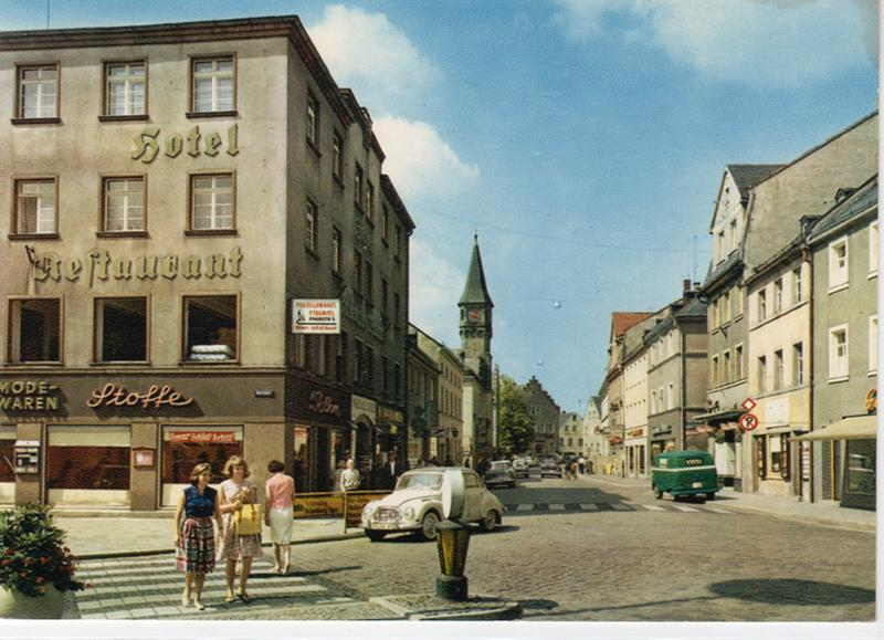 Farbige historische Innenstadtansicht mit Geschäften, Passanten, Fahrzeugen und der Stadtkirche im Hintergrund.