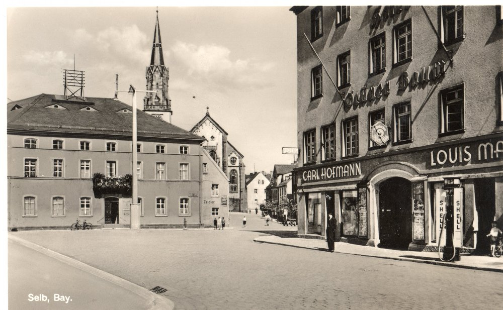 Historische Schwarzweißansicht des Selber Marktplatzes mit Ladengeschäften und der Stadtkirche im Hintergrund.
