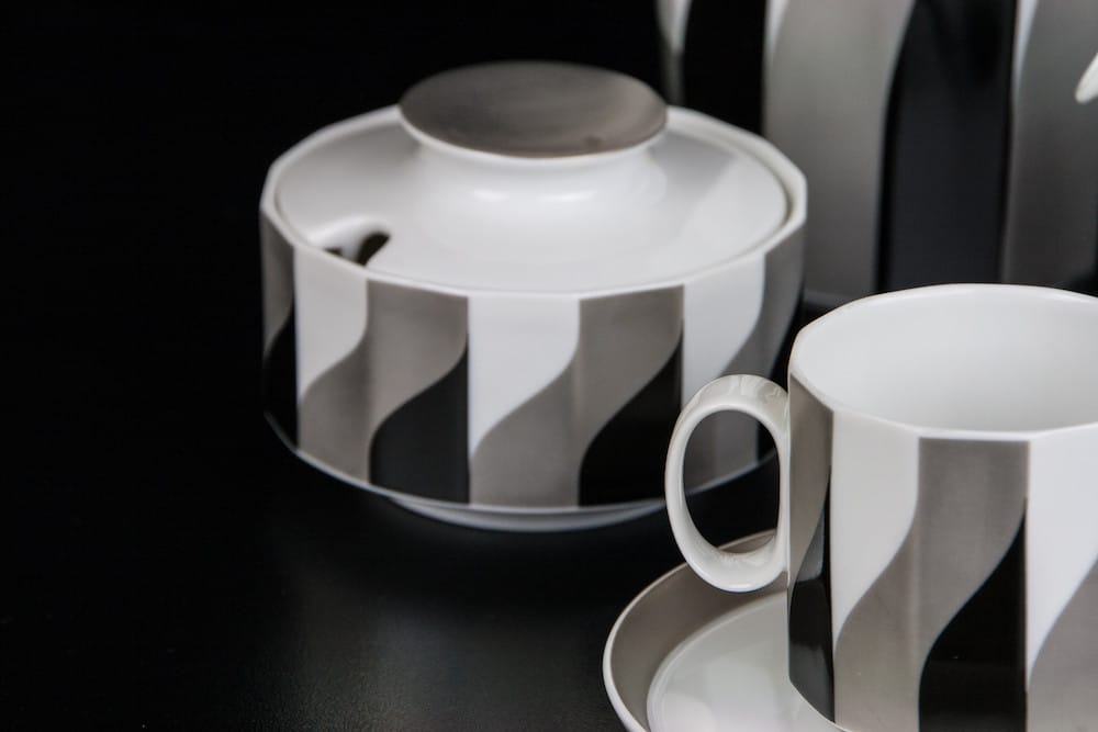 Weiße Kanne mit grauschwarzem flächigem Dekor neben einer weißen Tasse.