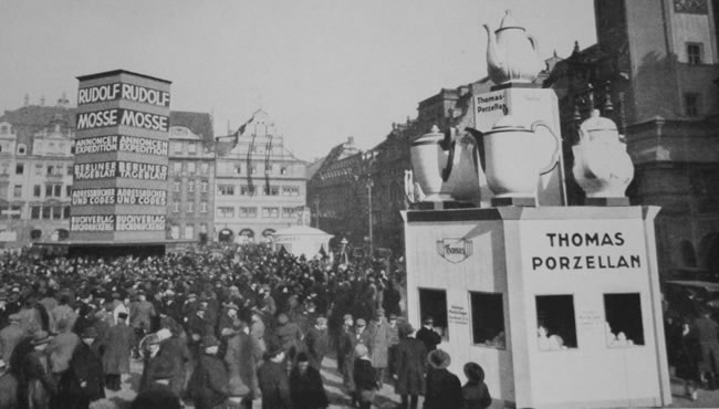 Historische Menschenmenge vor einer Thomas-Werbeanlage mit übergroßen Porzellankannen.