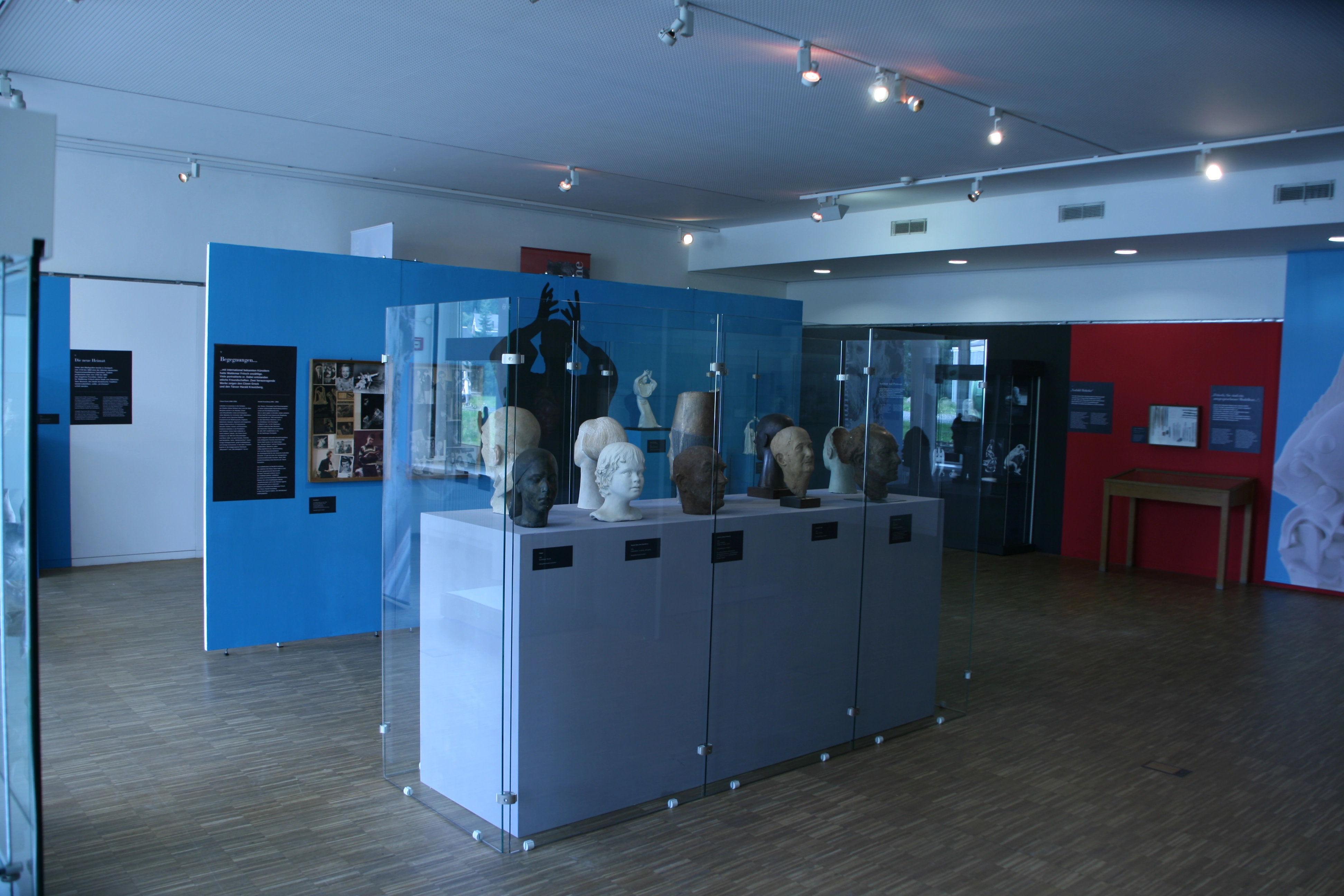 Blick in einen Ausstellungsraum mit Porzellanfiguren, Vitrinen und blauen sowie roten Stellwänden.