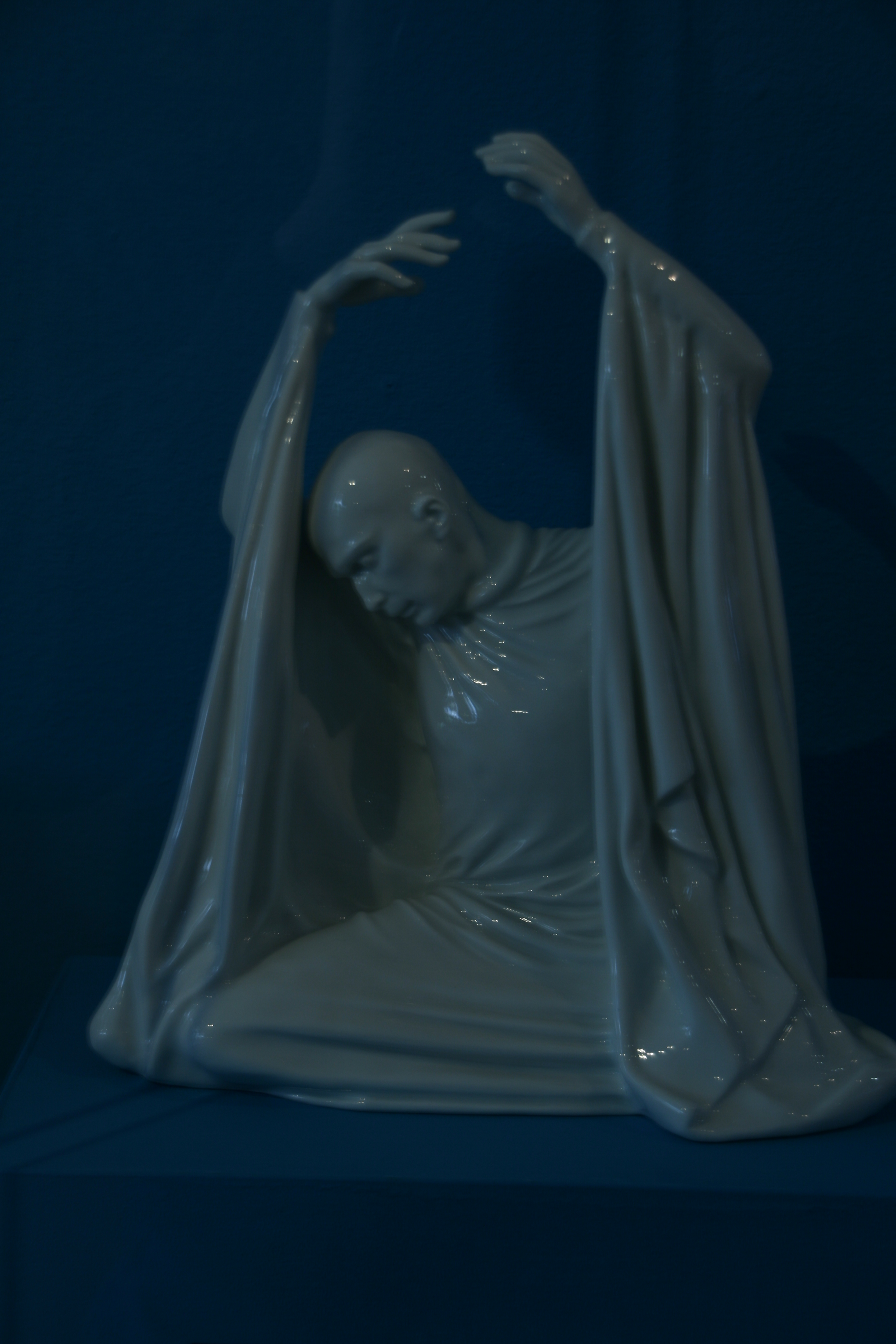 Weiße sitzende Porzellanfigur mit erhobenen Armen, gesenktem Kopf und bogenförmig fallenden Gewandteilen.