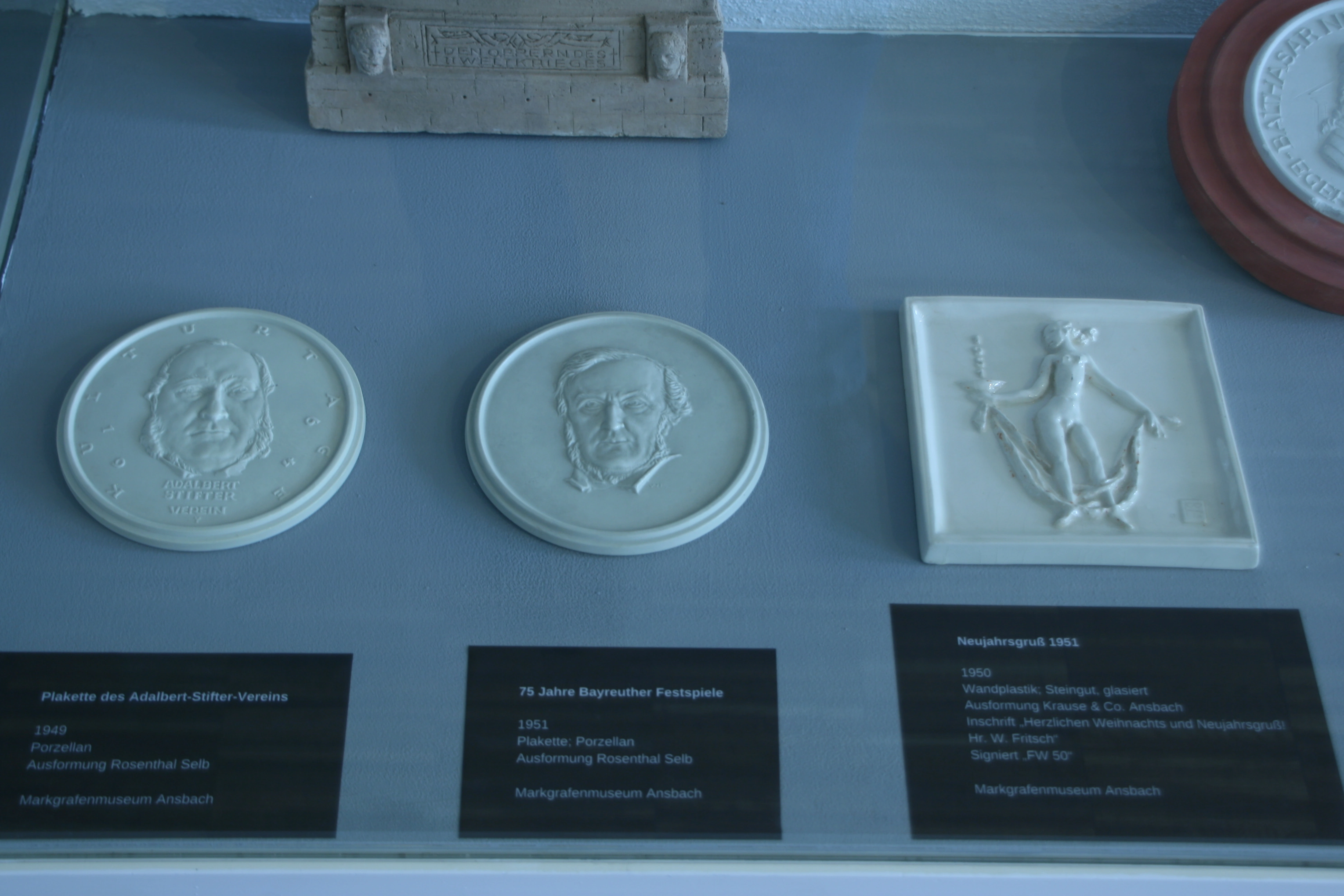 Zwei runde weiße Porträtplaketten und ein rechteckiges Relief; darunter die Museumsschilder.