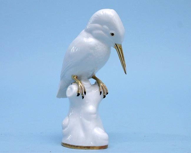 Weißer Porzellanvogel mit langem gelbem Schnabel und nach unten geneigtem Kopf auf einem hellen Astsockel.