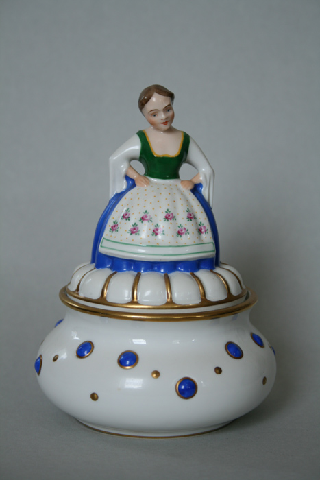 Runde weiße Porzellandose mit blauen Punkten und einer farbig bemalten Frauenfigur in Tracht auf dem Deckel.